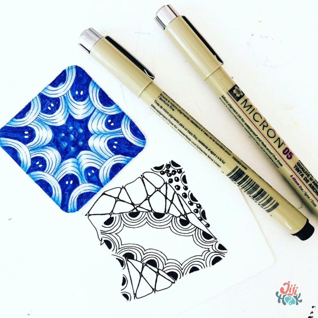 Mini tuile Zentangle bleue avec le motif Crescent Moon et un petit carré Zentangle en cours de dessin à côté de deux feutres de précision Sakura Micron